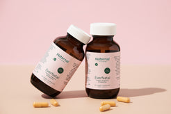 Two amber bottles of Naternal Vitamins EverNatal, a prenatal, pregnancy, and postpartum supplement, positioned on a beige surface with a soft pink background. The bottles, featuring white caps and minimalist labels, are surrounded by a few yellow capsules, highlighting maternal wellness support.