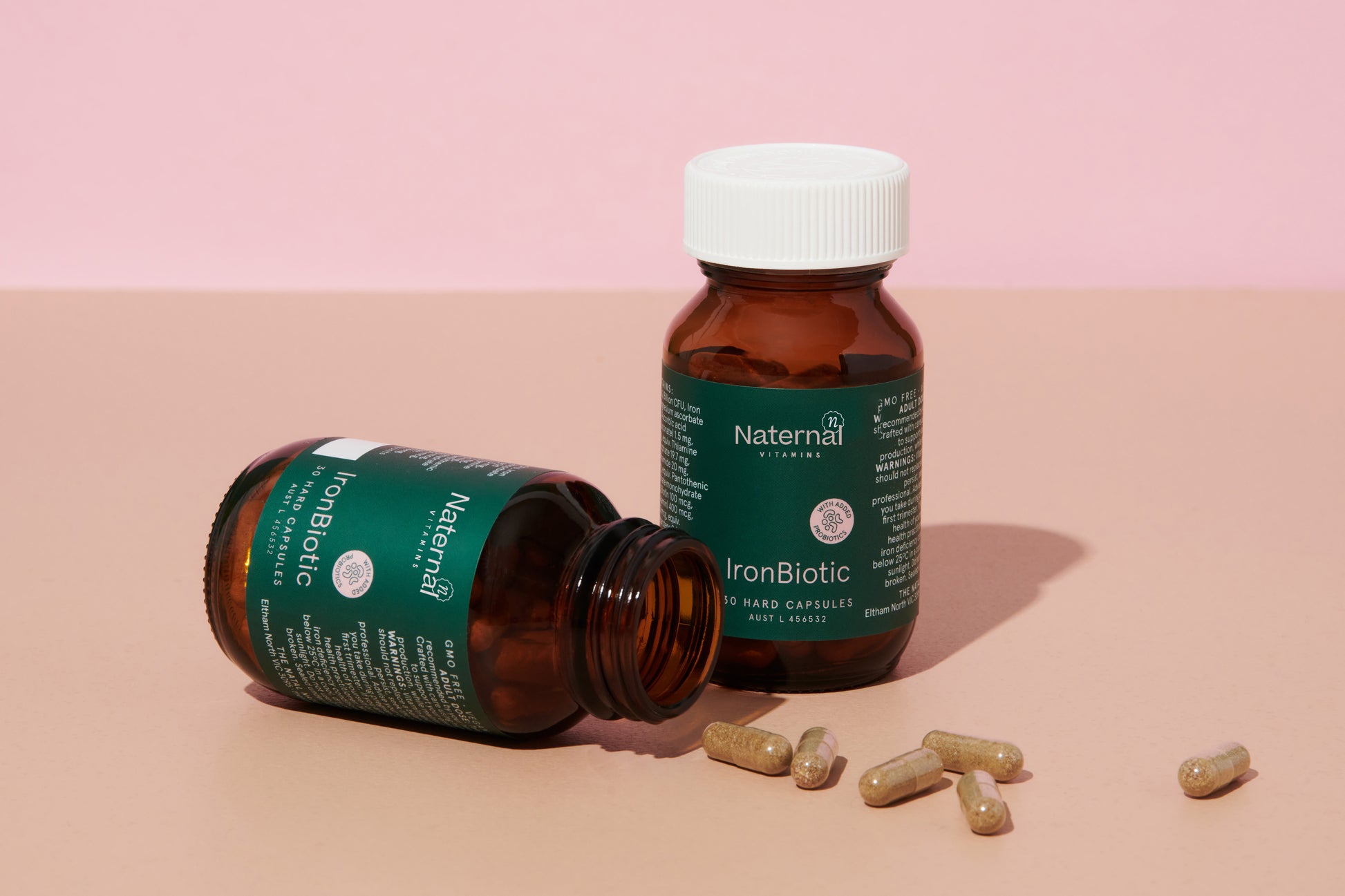 Two bottles of Naternal Vitamins IronBiotic supplement, with one bottle standing upright and the other lying open, spilling capsules onto a beige surface against a pink background. The product’s green label and white cap highlight its focus on iron and gut health support.