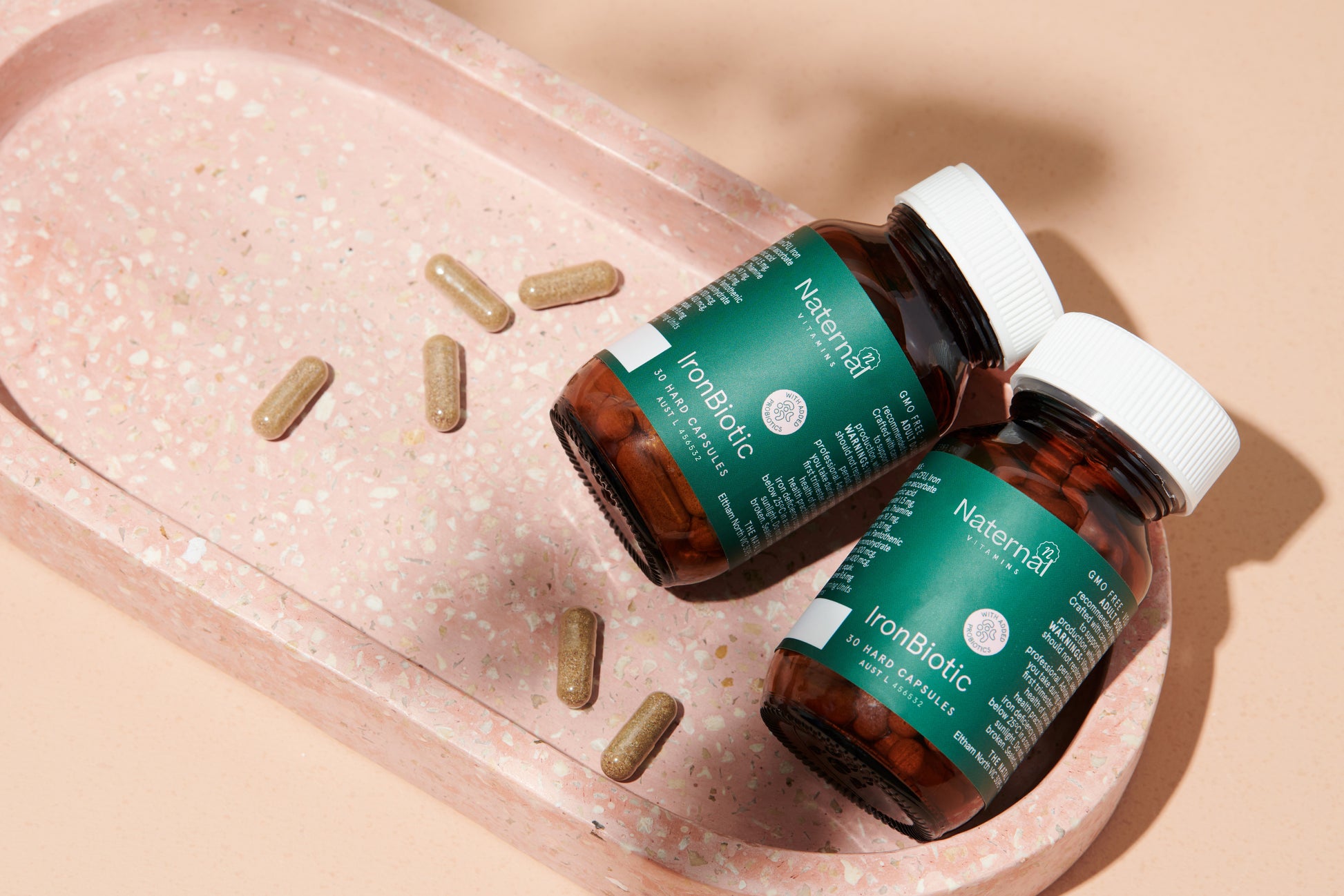 Two bottles of Naternal Vitamins IronBiotic, a blend designed to support iron levels and gut health, placed on a pink terrazzo tray with several capsules scattered around. The bottles, featuring a green label and white caps, convey a clean and health-focused aesthetic.