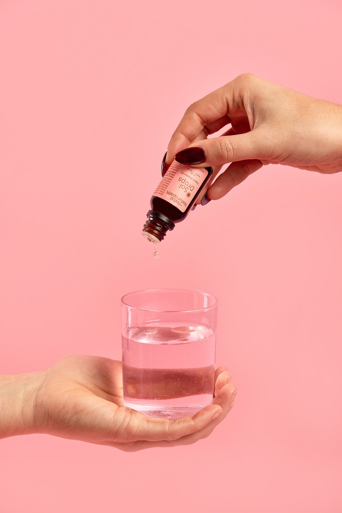 Hand pouring liquid from a dropper bottle into a glass on a pink background
