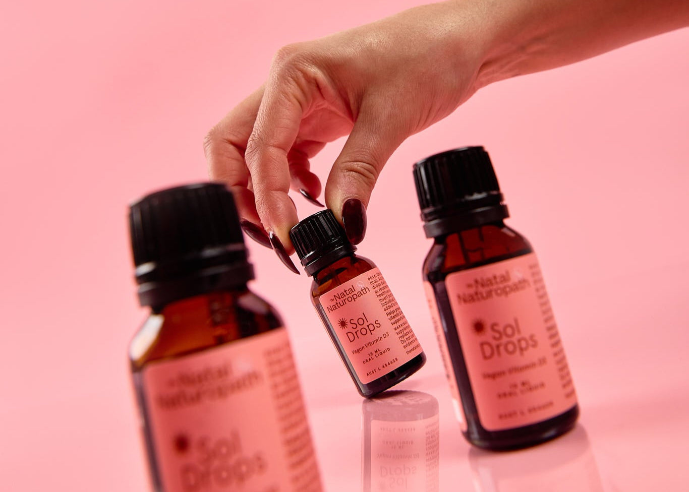 Hand holding three small bottles labeled 'Sol Drops' against a pink background