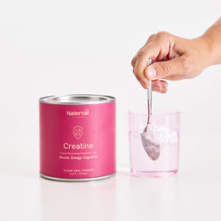 Person scooping creatine powder from a container into a glass with a spoon on a white background