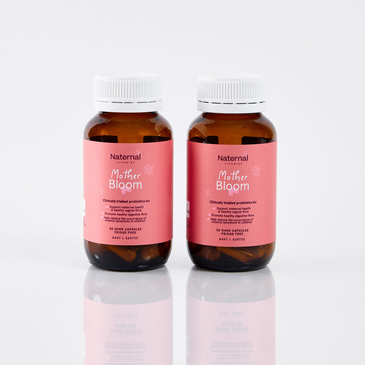 Two amber supplement bottles with pink labels reading 'Natural for Her Mother Bloom' and white ribbed caps, photographed on a clean white studio background with a soft reflection below.