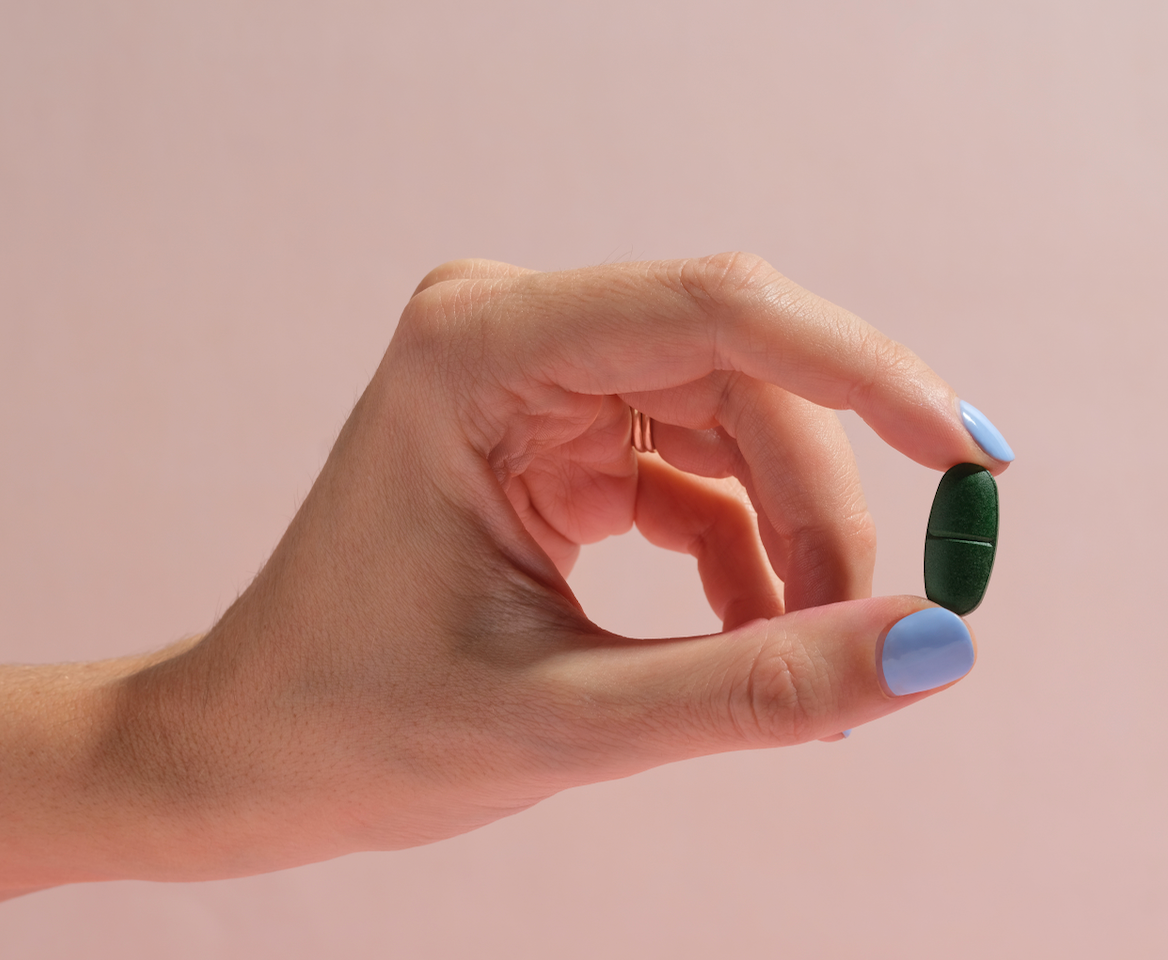 Close-up of a hand with blue-painted nails holding a single green tablet against a soft pink background. The minimalist composition highlights the tablet's focus on wellness and health support.