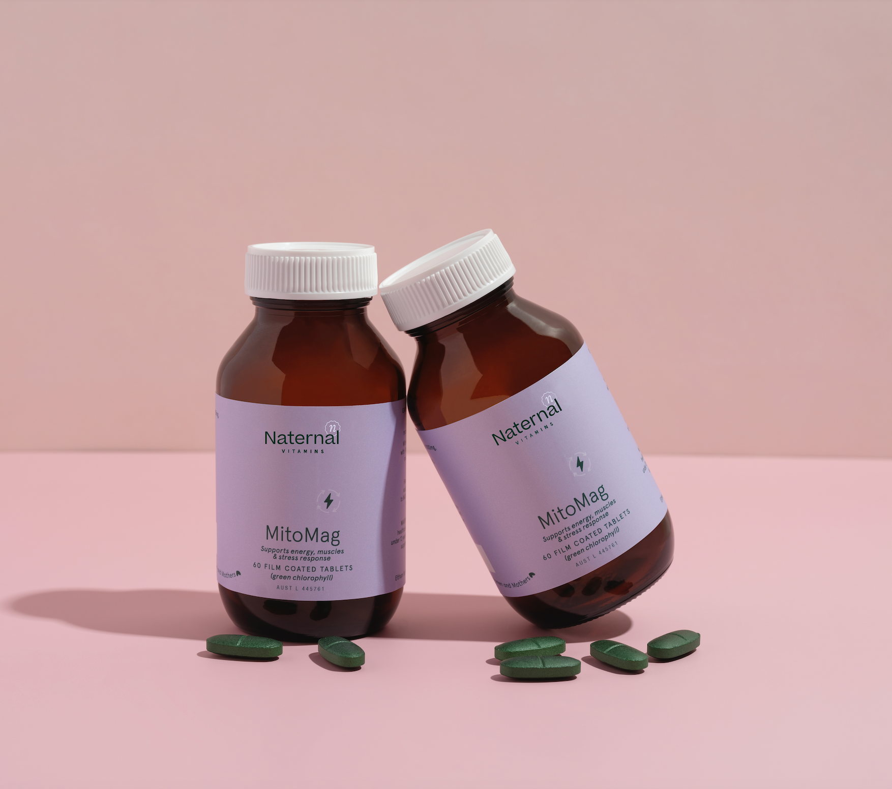 Two amber bottles of Naternal Vitamins MitoMag supplement with purple labels, placed on a pink surface against a soft background. The bottles are tilted toward each other, surrounded by several green tablets, highlighting the product's focus on supporting energy, muscle health, and stress response.