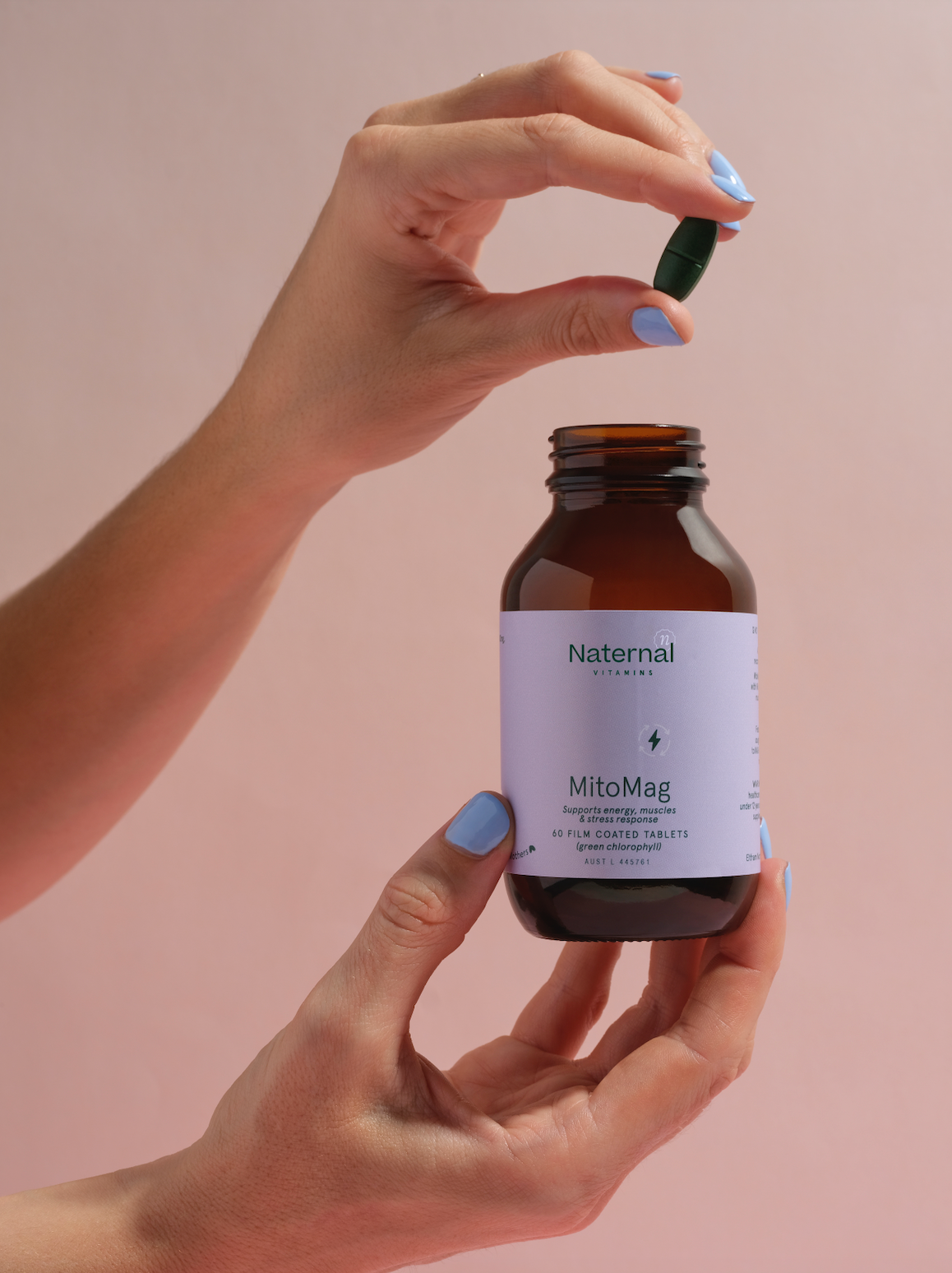 Close-up of hands holding a bottle of Naternal Vitamins MitoMag, a supplement supporting energy, muscle, and stress response. One hand holds the amber bottle with a soft purple label, while the other hand lifts a green tablet from the bottle, emphasising wellness and energy support.