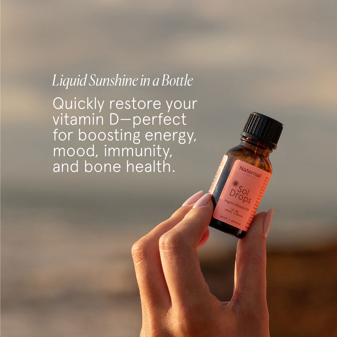 Hand holding a bottle labeled 'Sol Drops' with text about vitamin D benefits against a blurred natural background.