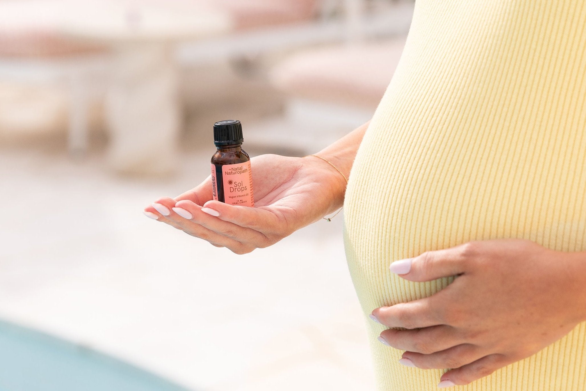 Close-up of a pregnant woman holding a bottle of Naternal Vitamins Sol Drops in one hand while gently cradling her belly with the other. Dressed in a soft yellow ribbed dress, the scene highlights a nurturing moment focused on wellness and prenatal care.