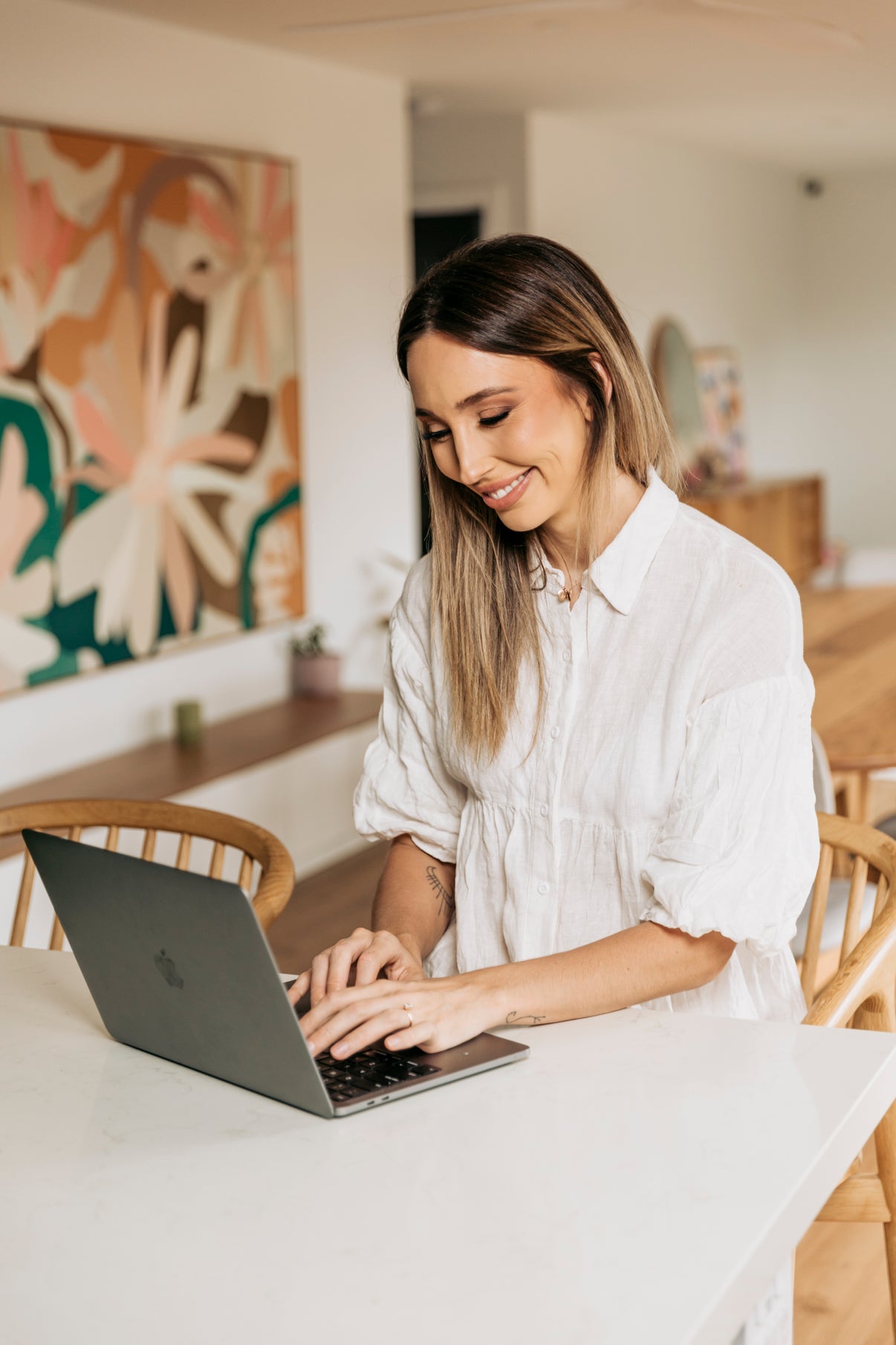 Melanie Nolan, founder of Naternal Vitamins, smiling while working on her laptop in a warmly decorated room with a botanical artwork in the background.